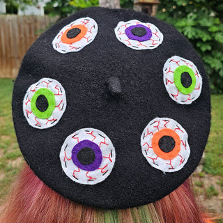 A black beret with a felt accent featuring multiple vibrant, hand-sewn eyeball designs on the front.